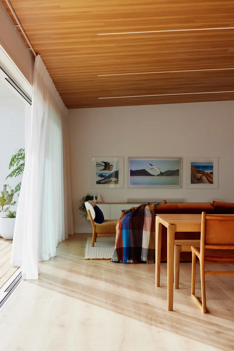 Open-plan living and dining space with sloped timber ceiling and floor-to-ceiling curtained glazing