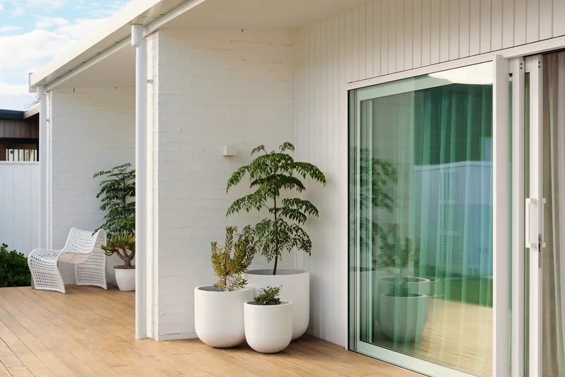 Covered timber deck with white painted brick wall, sliding glass door, and potted plants in planters