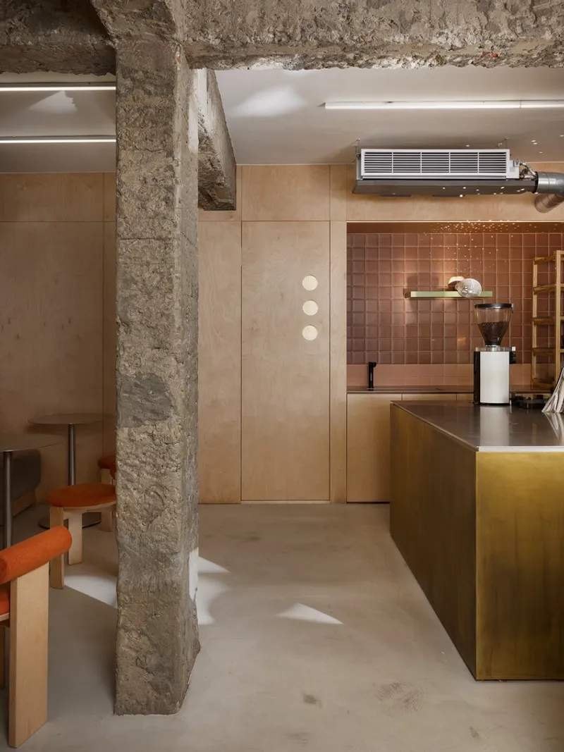 View past rough concrete column toward plywood cabinetry and terracotta tile kitchen