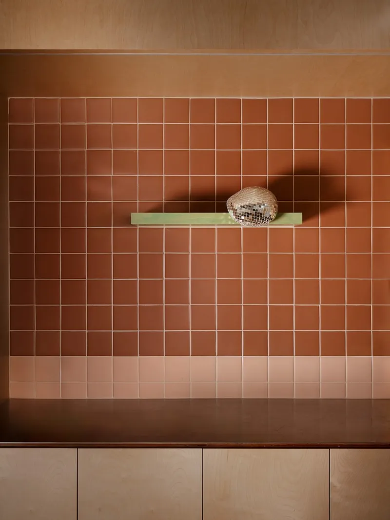 Gradient terracotta tile wall with green floating shelf displaying a mirrored disco ball under timber ceiling