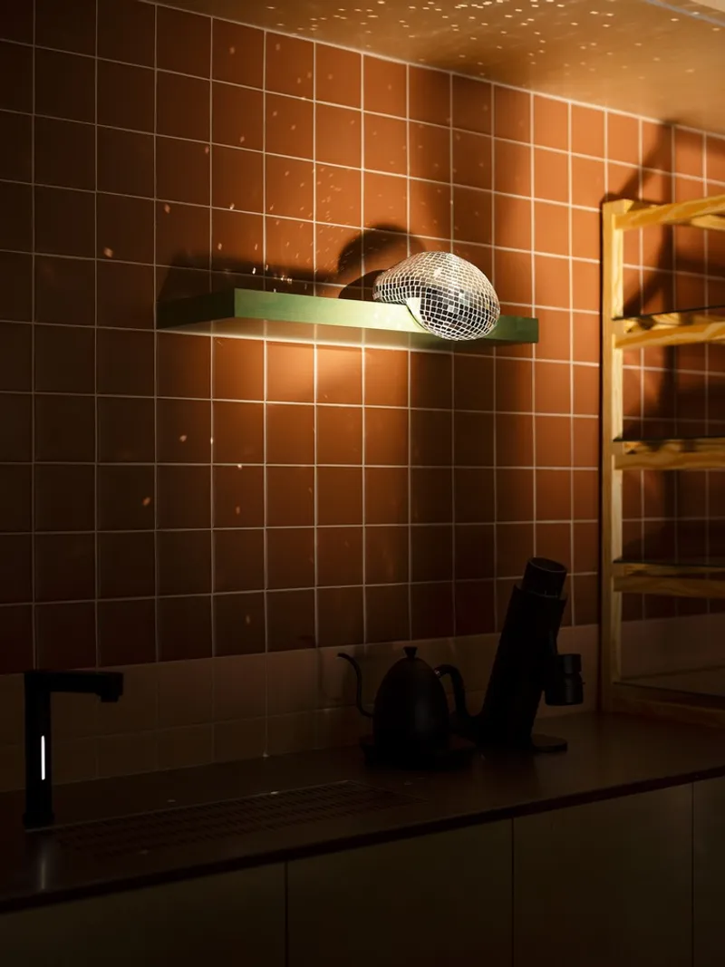 Kitchen counter with kettle beneath gradient terracotta tiled backsplash and green shelf in evening light