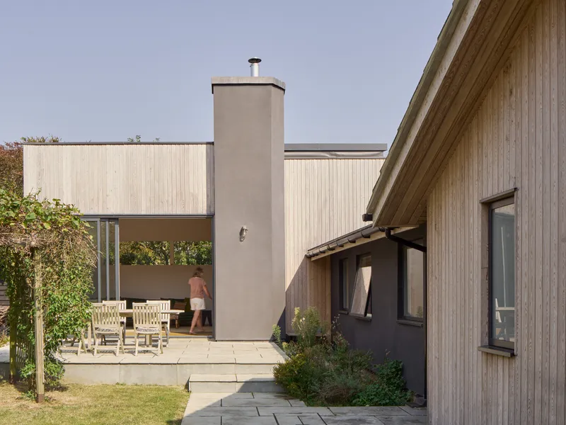 Courtyard terrace with concrete chimney, timber-clad volumes and person walking beyond dining furniture