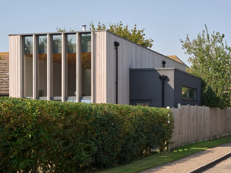 Street view showing timber-clad roof forms with vertical glazing above hedge and fence