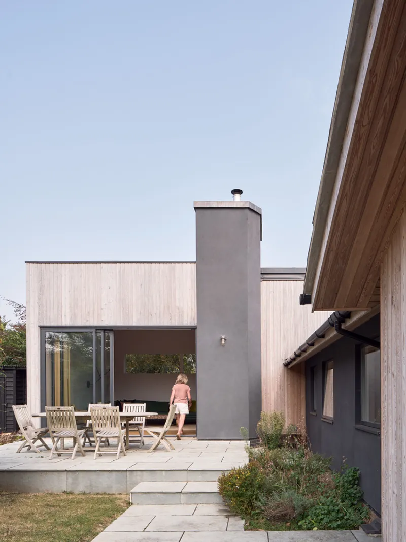Rear courtyard with concrete terrace, grey chimney stack and timber-clad volume with a person walking