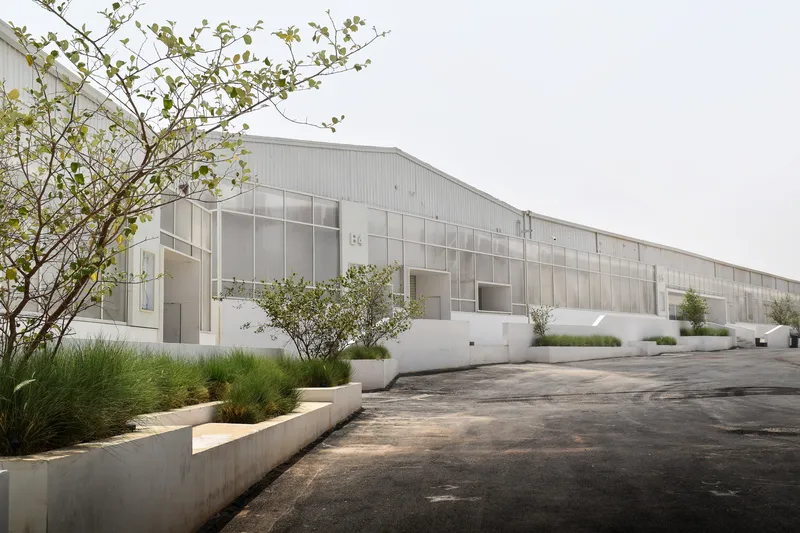 Long facade with translucent panels and concrete planters with grasses along a worn asphalt road