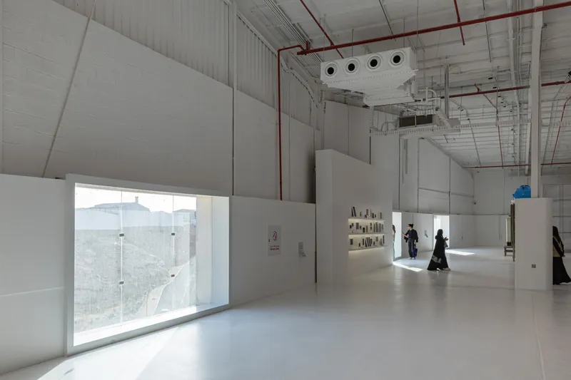 Interior gallery space with white partition walls, exposed ceiling ducts, and visitors in traditional dress