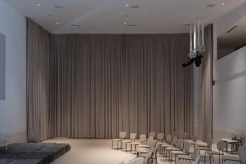 Interior performance space with floor-to-ceiling grey curtains and rows of chairs facing a stage