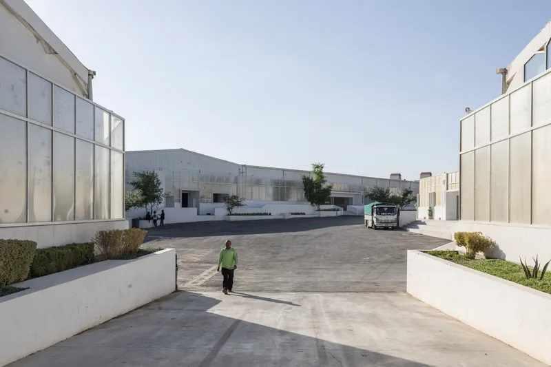 Paved courtyard with a worker walking toward metal-clad warehouse volumes and parked truck