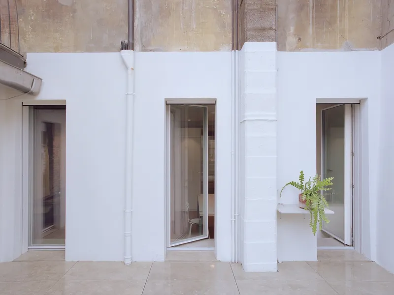 White courtyard wall with three tall windows framed in steel and exposed original masonry above