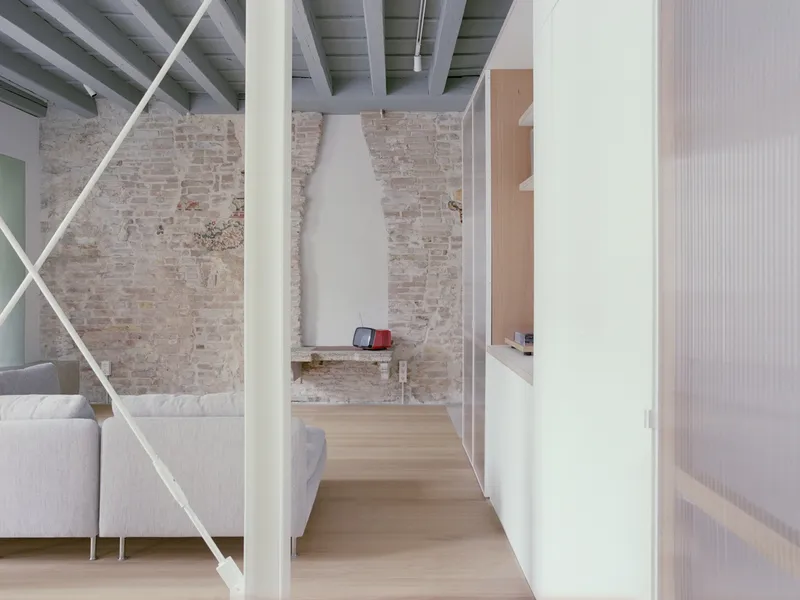 Interior view through a white steel-framed partition toward exposed brick walls and painted timber ceiling beams