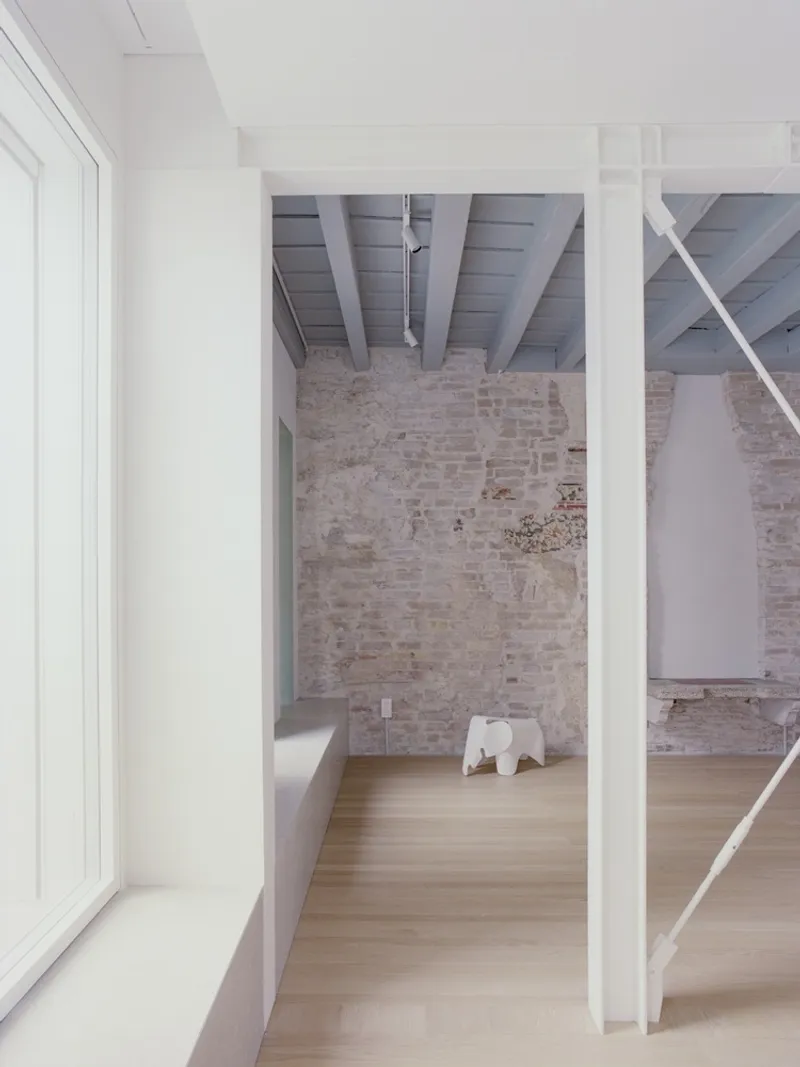 Corridor framed by white partition walls leading to a double-height space with exposed brick and painted beams