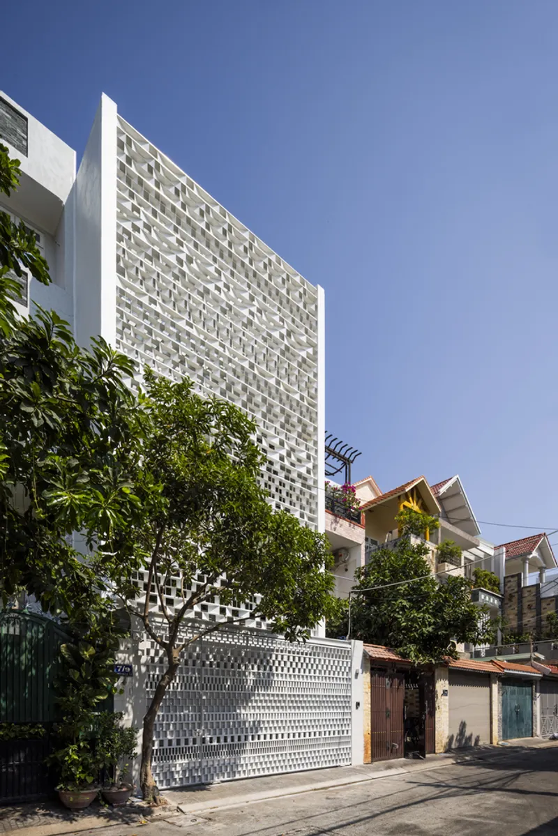 Street view of the perforated white facade rising above mature trees and a patterned boundary wall