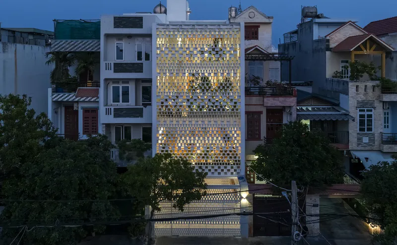 Perforated ceramic block facade glowing from interior lighting at dusk among neighboring houses