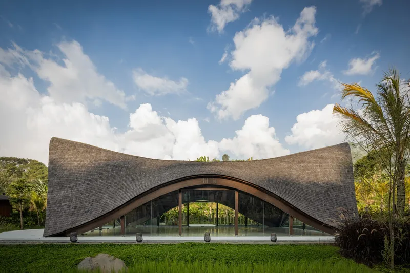 Arched pavilion with sweeping shingle roof and glass facade beneath billowing white clouds