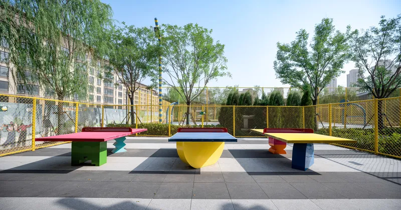 Painted benches in green, blue, and yellow arranged on grey paving with trees beyond yellow fence