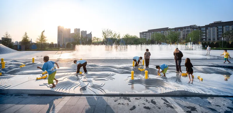 Interactive water fountain plaza with curved paving pattern and children playing at sunset
