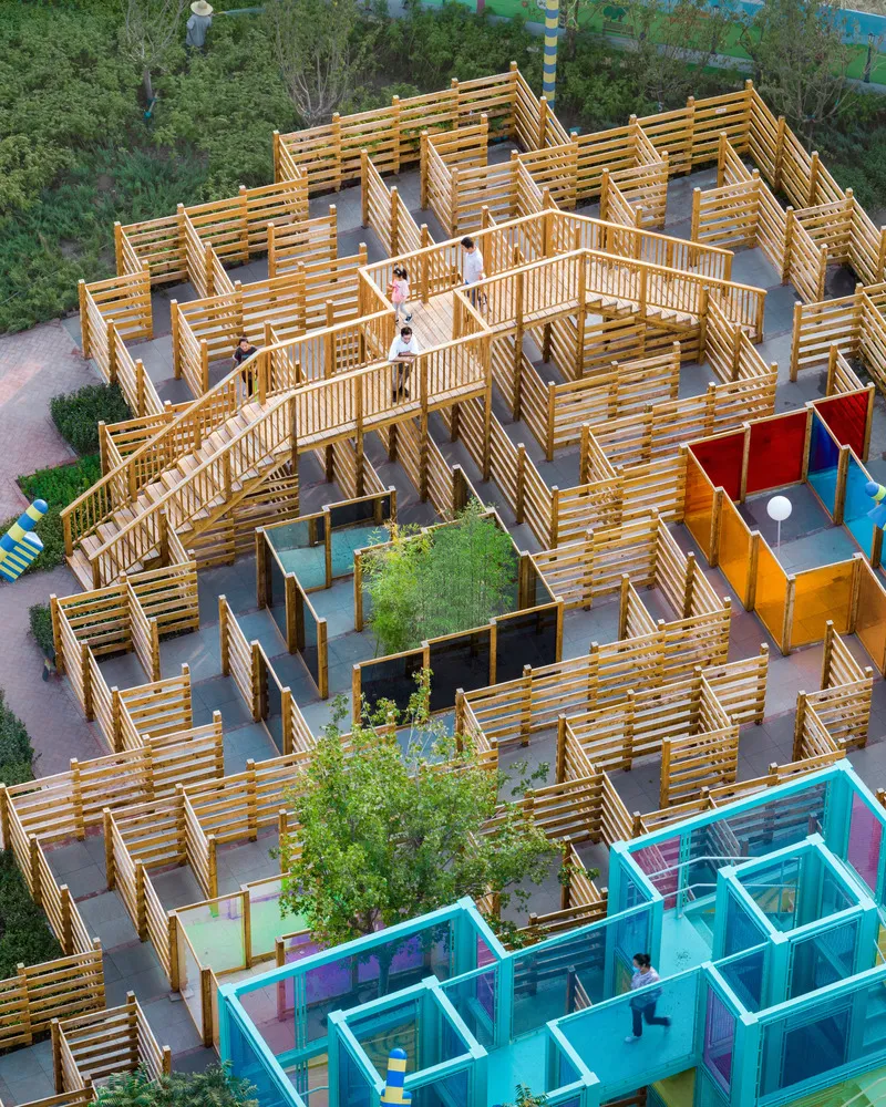 Wooden slatted maze structure with elevated walkways above colored cube pavilions and plantings