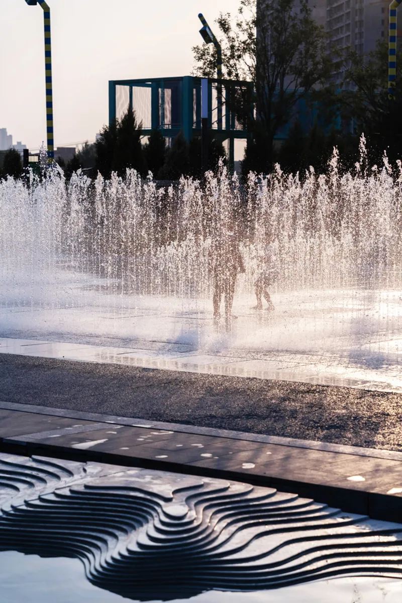 Fountain jets backlit at dusk with sculptural metal bench and building frames beyond