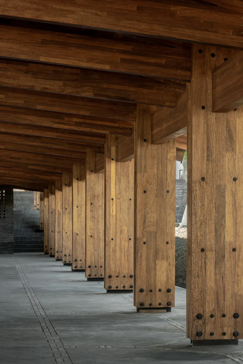 Covered corridor with timber columns and beams casting shadows across a polished concrete floor
