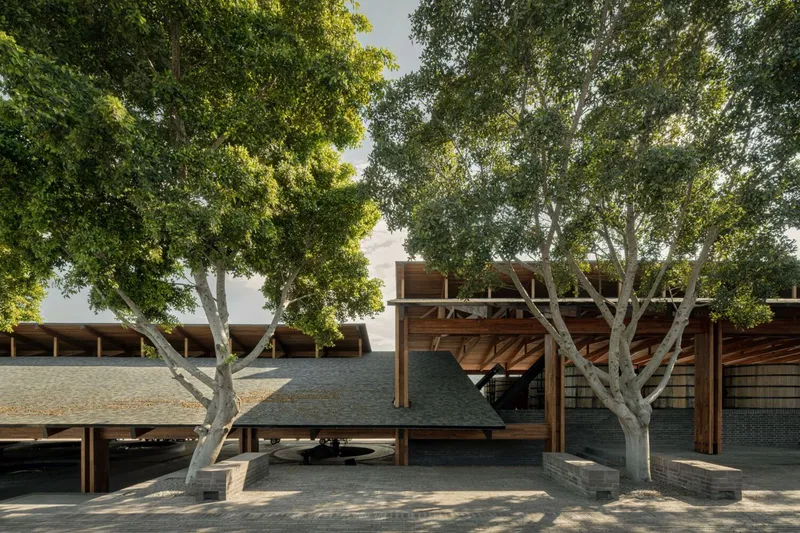 Brick and timber pavilion with exposed rafters framed by mature deciduous trees in dappled sunlight