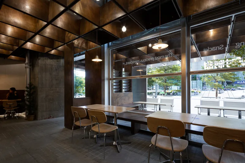 Communal seating area with wooden chairs beneath coffered ceiling and full-height glazing overlooking street trees