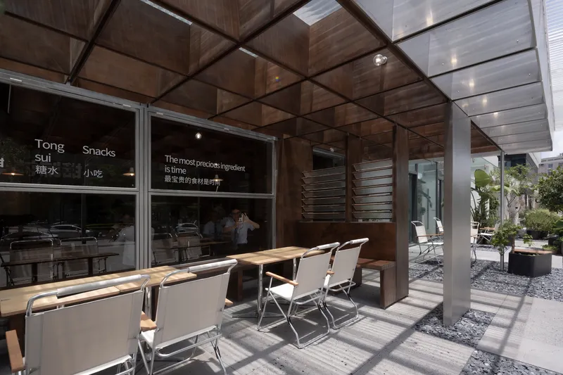 Outdoor seating terrace with folding chairs under coffered overhang casting dappled shadow on concrete pavers