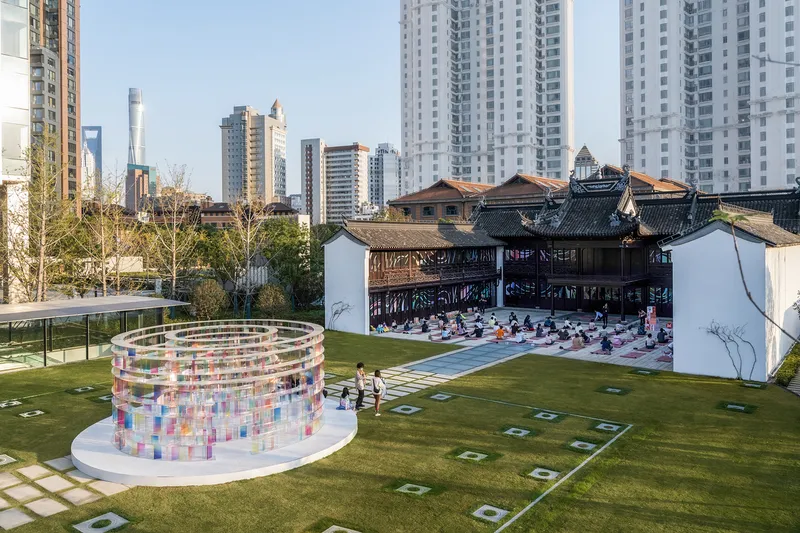 Circular pavilion with translucent colored panels on a lawn beside a traditional timber building