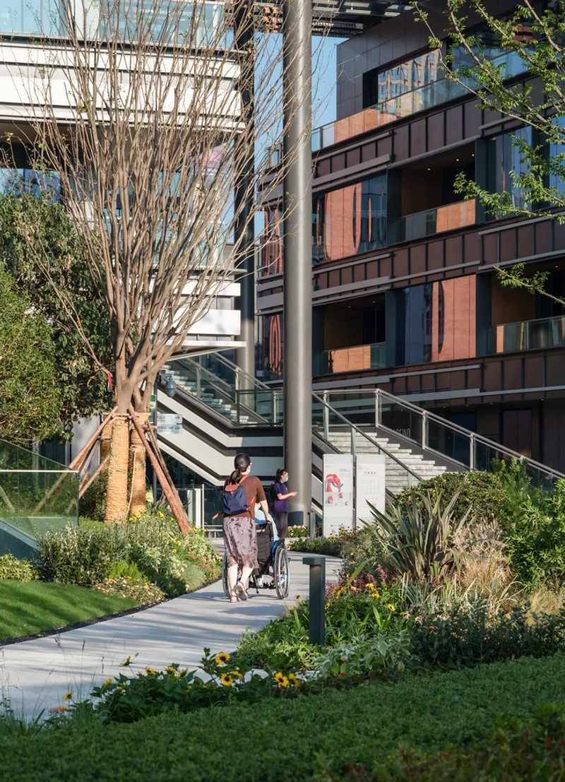Landscaped walkway with visitors and a stroller passing between planted beds and a copper-paneled facade