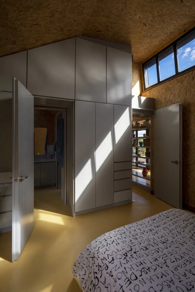 Bedroom interior with pale grey wardrobe unit, OSB walls, high clerestory windows, and yellow polished floor