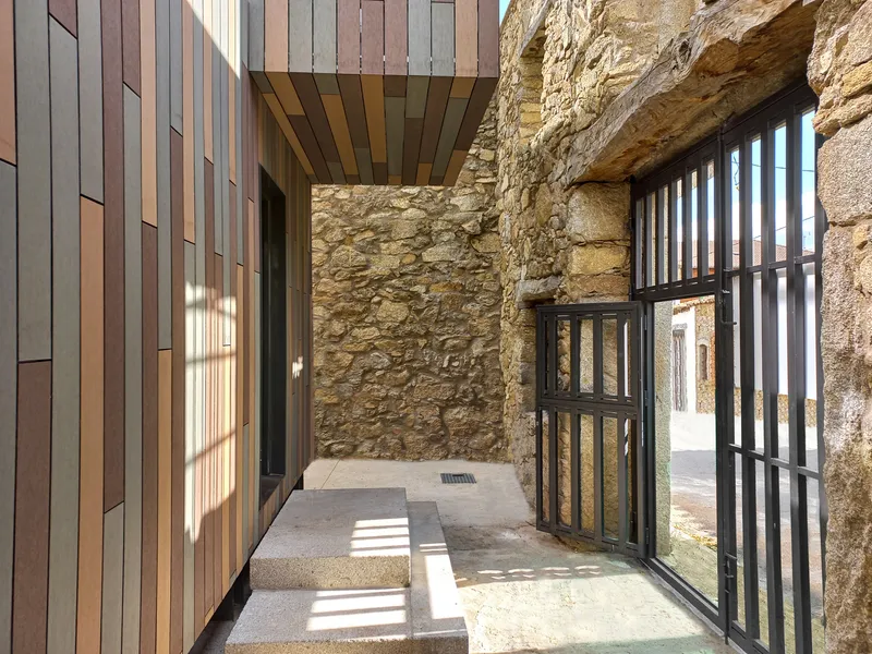 Entry passage between timber-clad volume and stone wall with metal-framed gate filtering afternoon light
