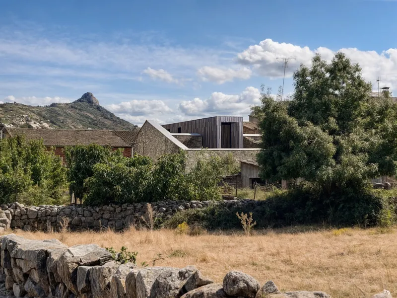 Distant view of the timber-clad volume rising above surrounding stone walls and trees with hillside beyond