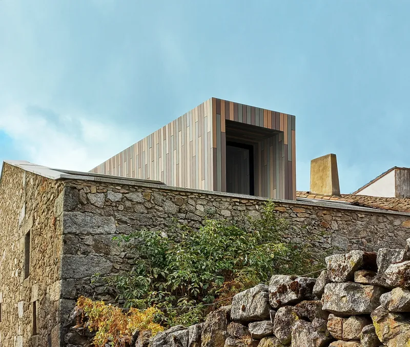 Upper volume clad in variegated timber planks projecting above weathered stone walls and low vegetation