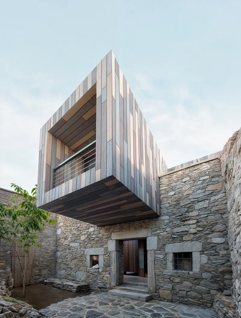 Cantilevered timber-clad volume suspended above the stone courtyard entry with exposed soffit and planted tree