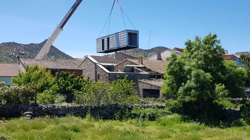 Prefabricated module suspended by crane above stone houses with green trees and hillside backdrop