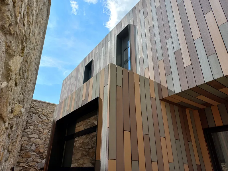 Facade of vertical metal panels in bronze and grey tones beside weathered stone wall