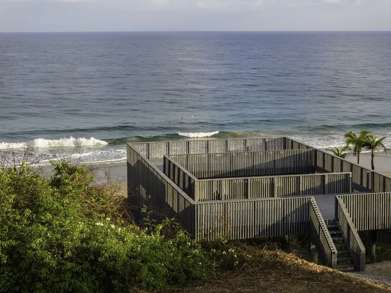 Elevated timber structure with slatted facade and staircase facing the surf and palm trees