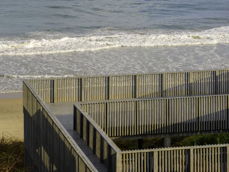 Wooden deck and stair descending from slatted facades toward the breaking waves on the beach