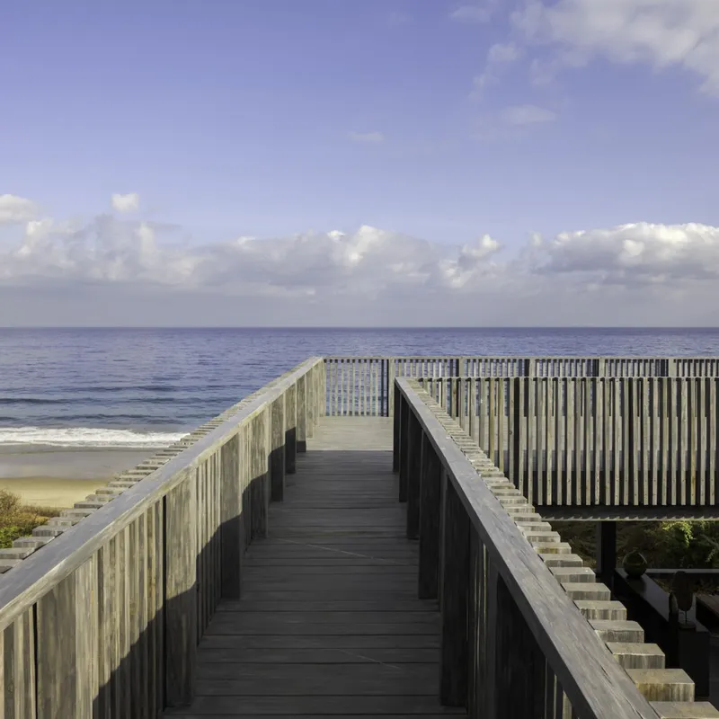 Timber boardwalk with vertical slat railings leading toward the ocean under scattered clouds