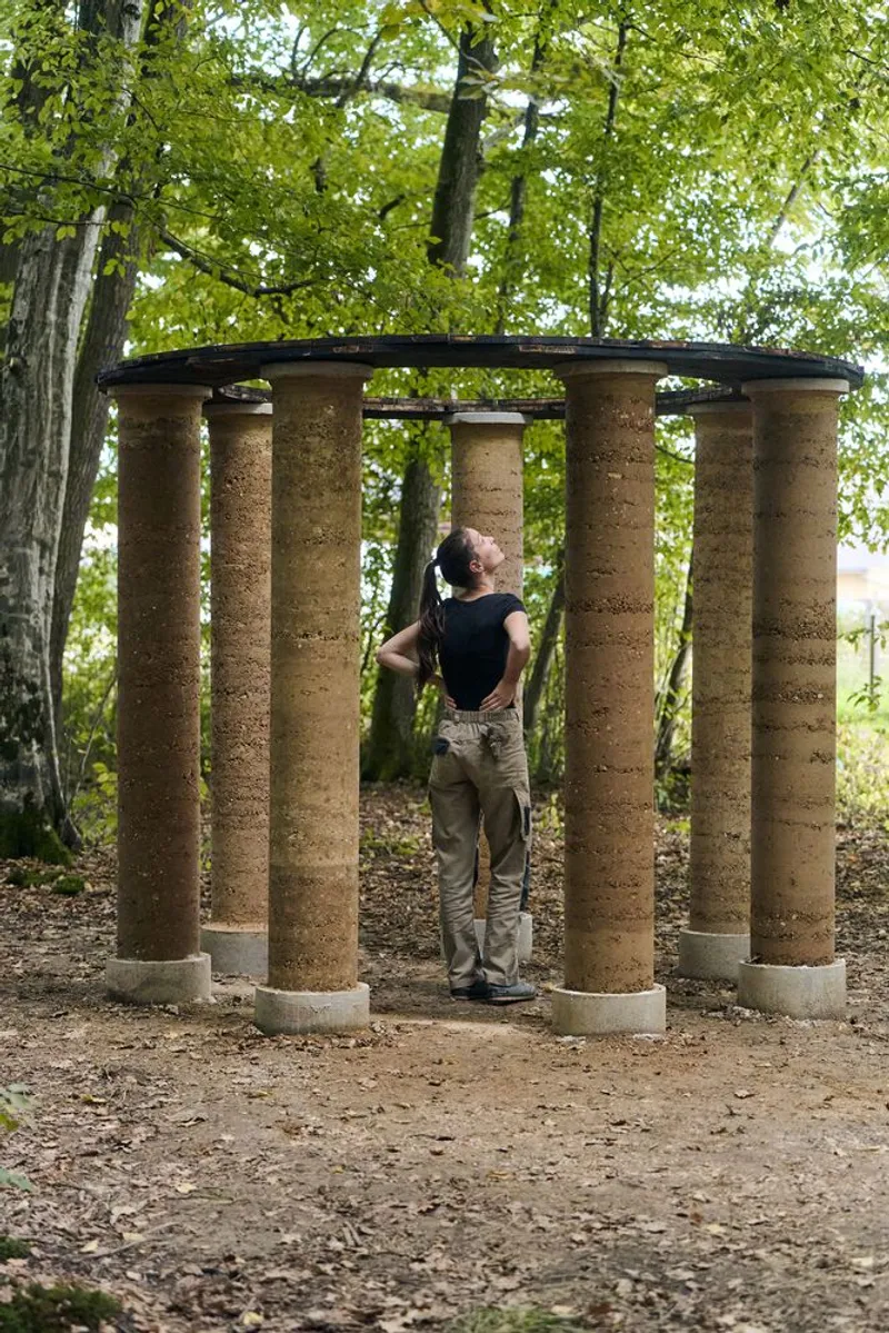 Circular colonnade of cylindrical rammed earth columns supporting a thin roof with visitor reaching upward
