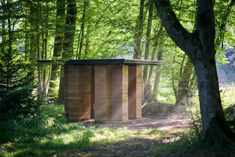Small rammed earth pavilion with flat roof nestled in a forest clearing with dappled sunlight