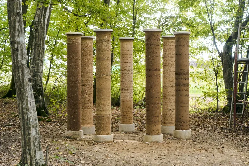 Ring of six rammed earth columns supporting a flat roof on a gravel clearing