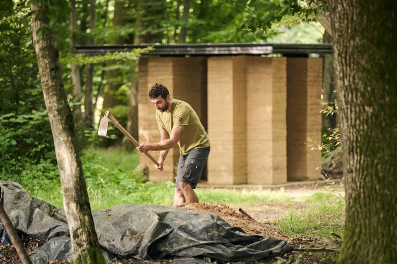 Man working with an axe in front of a small plywood structure in a wooded clearing
