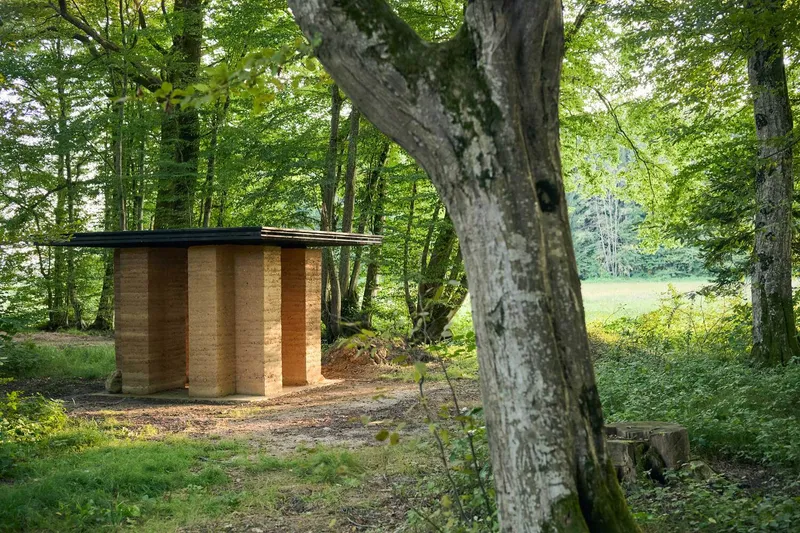 Small rammed earth pavilion with flat roof nestled among trees at the edge of a forest clearing