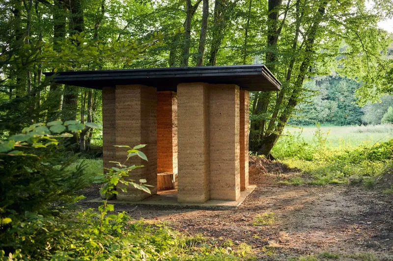 Open shelter with layered earth walls and overhanging roof framed by dappled sunlight through the canopy
