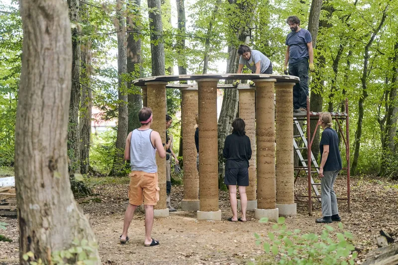 Construction team assembling the roof structure atop cylindrical rammed earth columns in a woodland setting