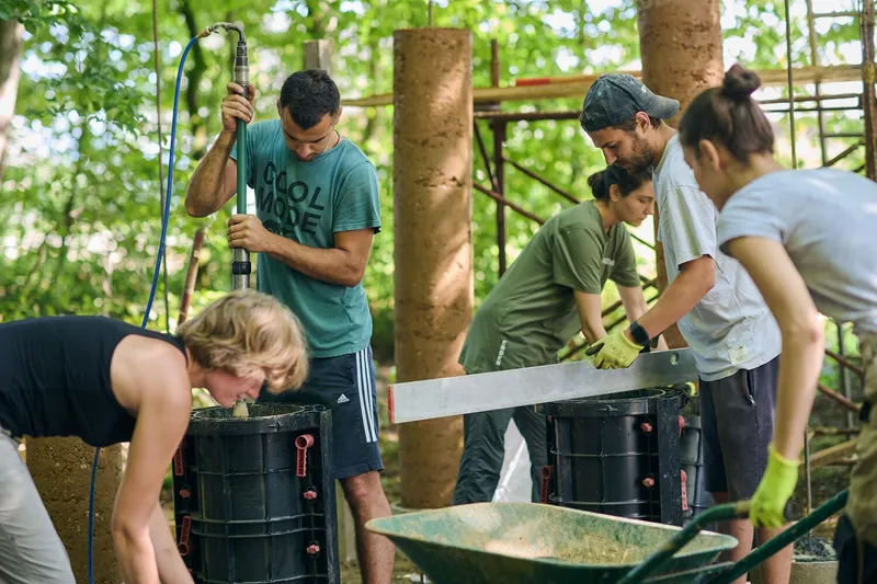 Group of builders tamping earth into cylindrical molds and mixing material in a wheelbarrow on site