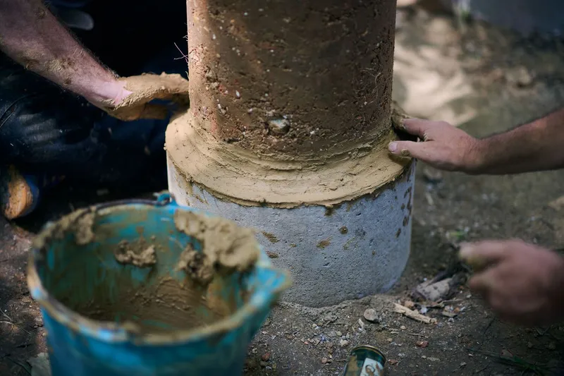 Hands applying earth plaster to the base of a rammed earth column beside a blue mixing bowl