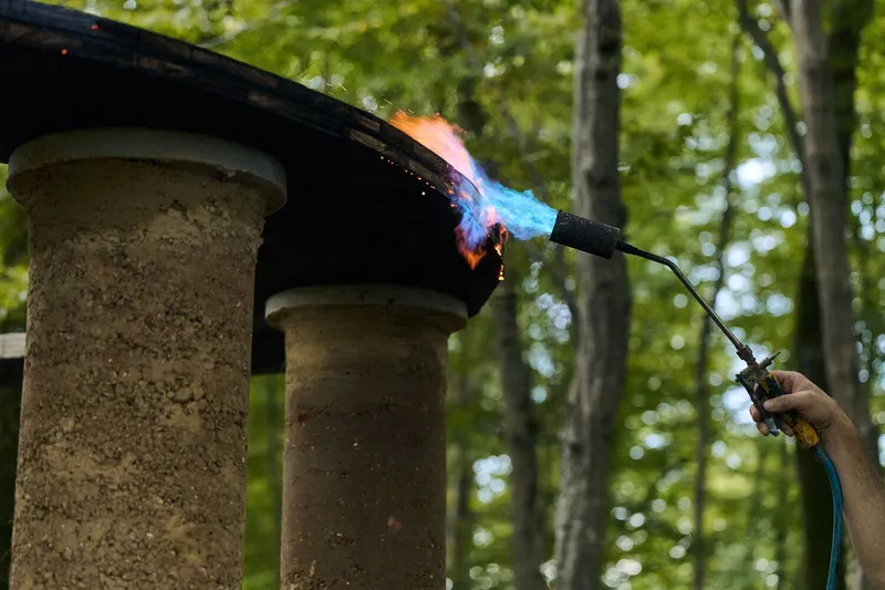 Gas torch flame treating the capital of a rammed earth column in a wooded setting