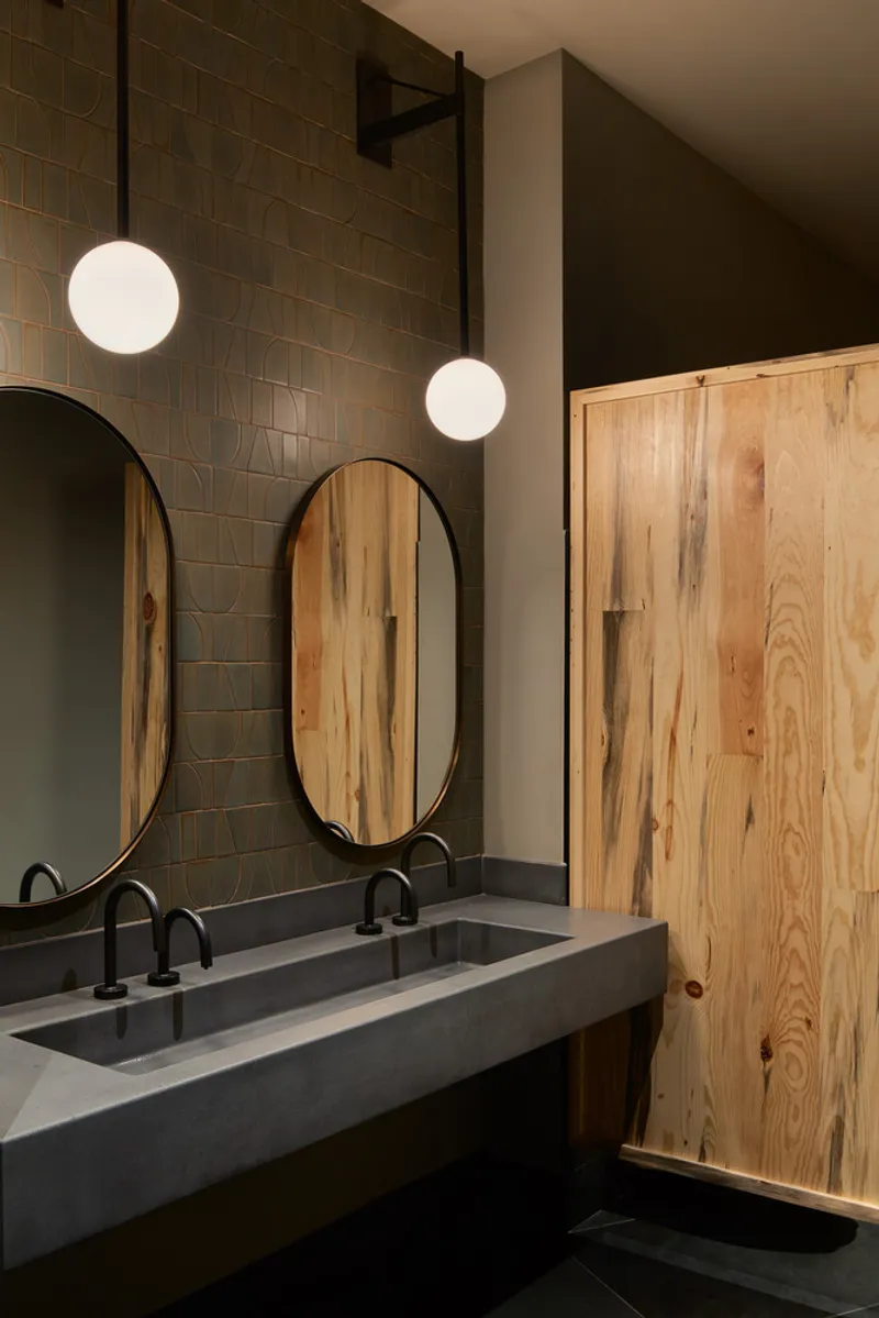 Washroom with concrete trough sink, oval mirrors and knotted timber partition under globe pendants