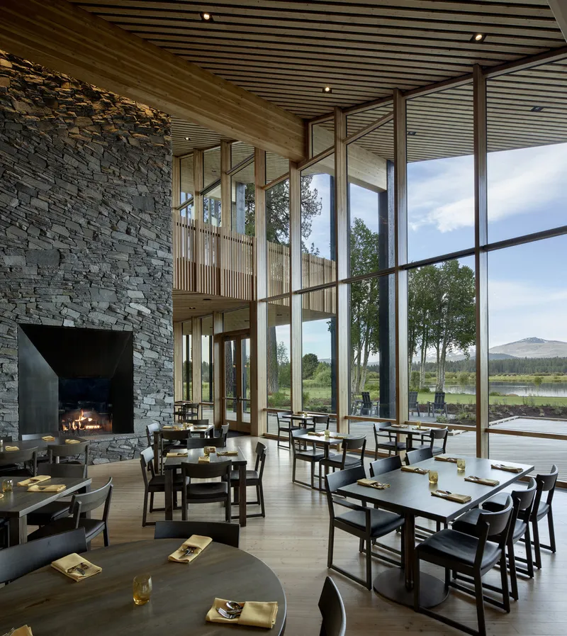Double-height dining hall with stacked stone fireplace, timber screens and floor-to-ceiling glazing overlooking a lake